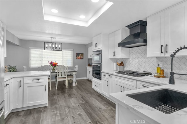 kitchen with multiple ovens, a tray ceiling, wood finished floors, wall chimney range hood, and stainless steel gas cooktop