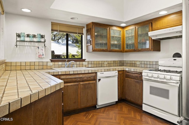 kitchen with under cabinet range hood, a sink, white appliances, glass insert cabinets, and tile counters