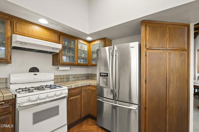 kitchen featuring tile countertops, white range with gas stovetop, stainless steel fridge, and under cabinet range hood