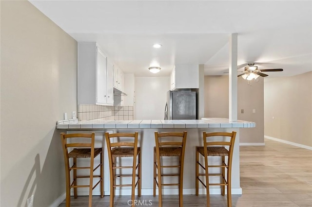 kitchen with backsplash, tile countertops, light wood-type flooring, a peninsula, and freestanding refrigerator