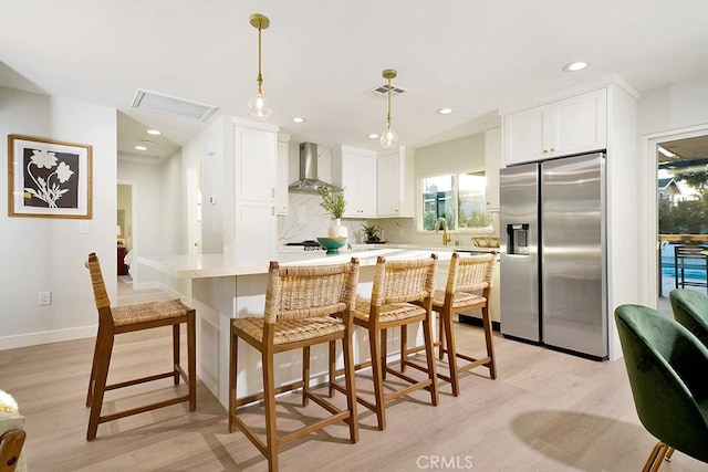 kitchen featuring visible vents, wall chimney exhaust hood, stainless steel refrigerator with ice dispenser, and white cabinets