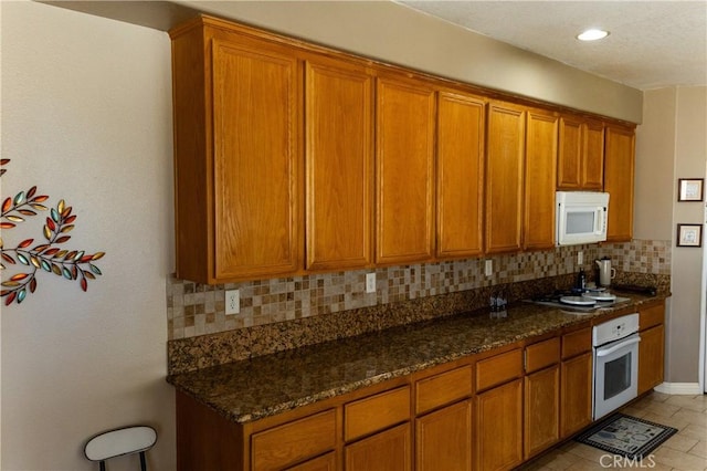 kitchen featuring backsplash, recessed lighting, dark stone countertops, brown cabinetry, and white appliances