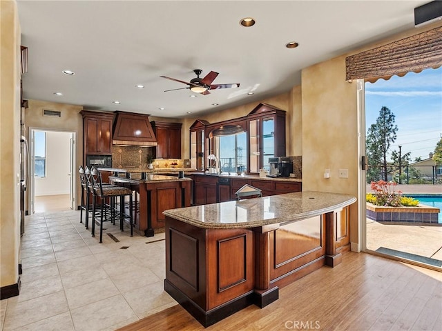 kitchen with visible vents, light stone countertops, premium range hood, decorative backsplash, and a kitchen breakfast bar