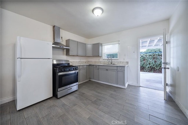 kitchen with freestanding refrigerator, a sink, gray cabinetry, wall chimney exhaust hood, and stainless steel gas stove