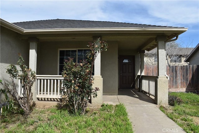 view of exterior entry with stucco siding, covered porch, a shingled roof, and fence