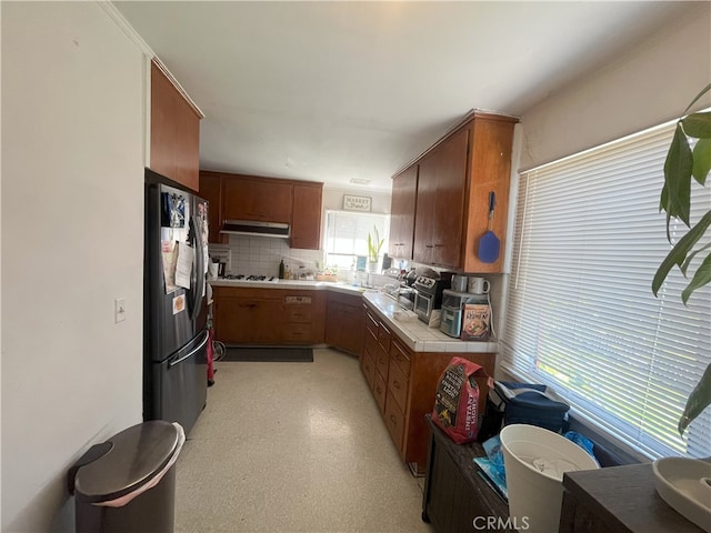 kitchen featuring brown cabinets, under cabinet range hood, backsplash, freestanding refrigerator, and tile counters