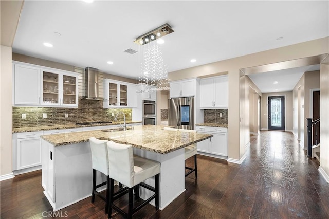 kitchen featuring a center island with sink, dark wood finished floors, a sink, appliances with stainless steel finishes, and wall chimney exhaust hood