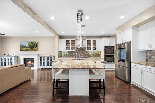 kitchen with a warm lit fireplace, dark wood-style floors, stainless steel appliances, wall chimney exhaust hood, and glass insert cabinets