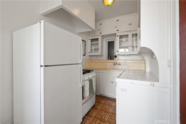 kitchen with tasteful backsplash, glass insert cabinets, tile countertops, white appliances, and white cabinetry