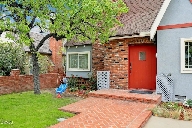 view of exterior entry with stucco siding, fence, a yard, a shingled roof, and brick siding