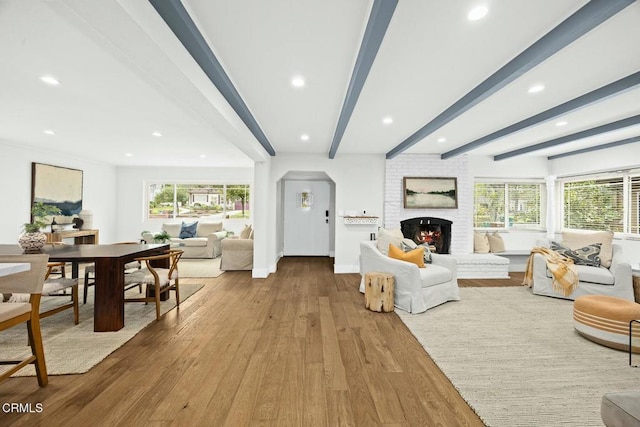 living area with a wealth of natural light, beam ceiling, a brick fireplace, and wood finished floors