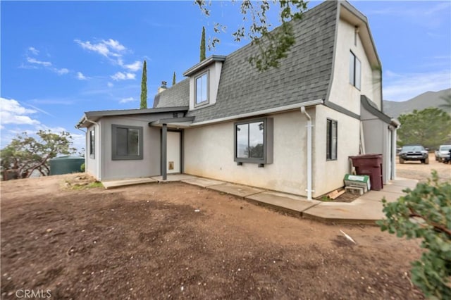 back of property with a shingled roof, mansard roof, stucco siding, a patio, and a mountain view