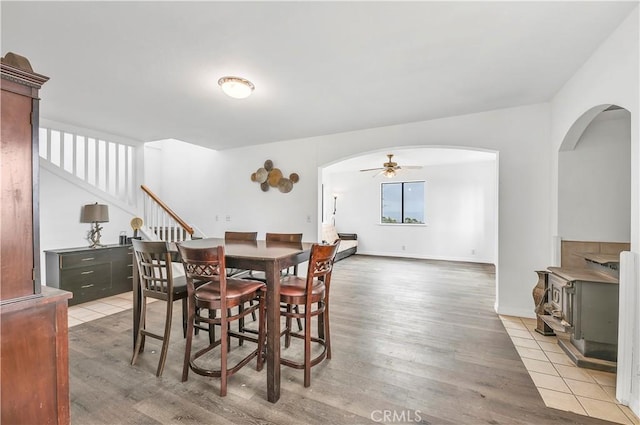 dining space with a wood stove, arched walkways, light wood-type flooring, stairs, and ceiling fan