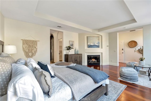 bedroom with wood finished floors, visible vents, baseboards, a tray ceiling, and a glass covered fireplace