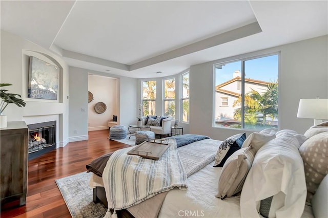 bedroom featuring a glass covered fireplace, a tray ceiling, baseboards, and wood finished floors