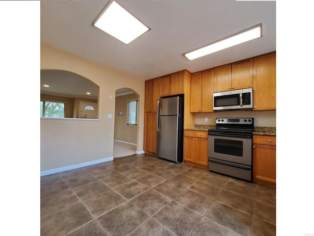 kitchen with baseboards, dark stone counters, dark tile patterned flooring, arched walkways, and stainless steel appliances