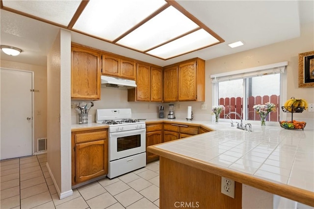 kitchen with white gas stove, a peninsula, under cabinet range hood, and tile counters