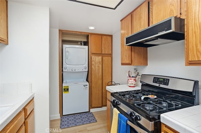 kitchen with tile counters, under cabinet range hood, gas range, light wood-type flooring, and stacked washer / drying machine