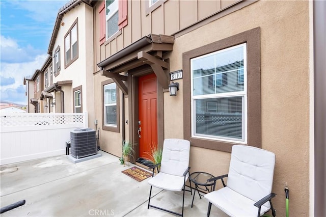 view of exterior entry featuring board and batten siding, fence, central air condition unit, stucco siding, and a patio