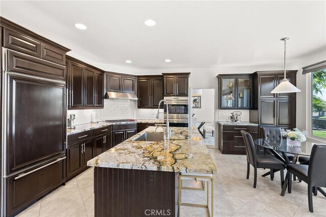 kitchen with light tile patterned flooring, hanging light fixtures, dark brown cabinetry, under cabinet range hood, and tasteful backsplash