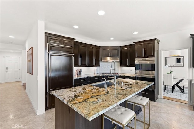 kitchen featuring under cabinet range hood, tasteful backsplash, a breakfast bar area, light stone countertops, and paneled built in refrigerator