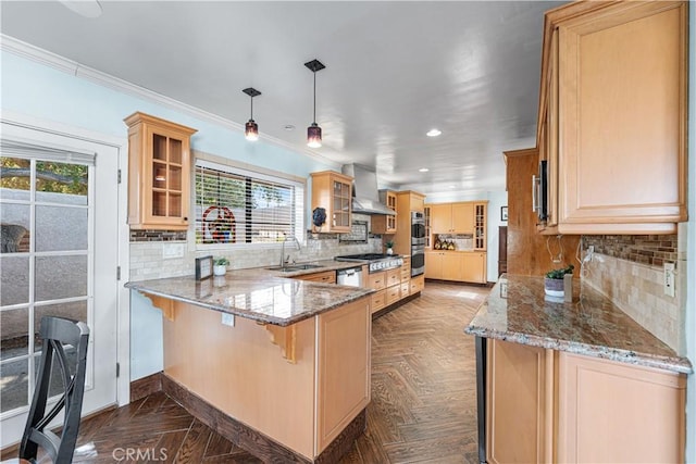 kitchen featuring a peninsula, light brown cabinetry, a sink, glass insert cabinets, and wall chimney exhaust hood