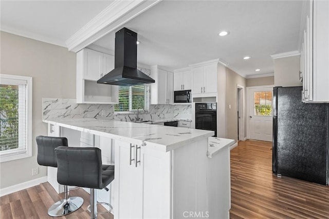 kitchen with light stone counters, a peninsula, black appliances, white cabinets, and island range hood