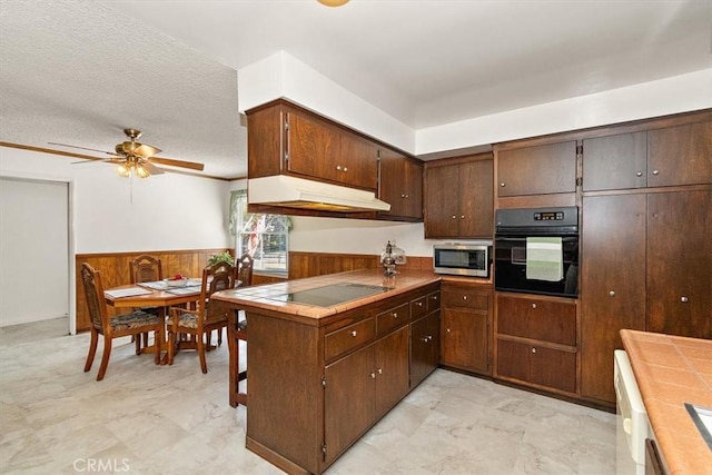 kitchen with a wainscoted wall, black appliances, under cabinet range hood, a textured ceiling, and wooden walls