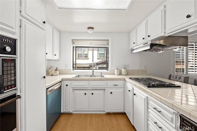 kitchen featuring stainless steel dishwasher, tile counters, under cabinet range hood, and a sink