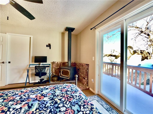 bedroom featuring access to exterior, ceiling fan, a wood stove, wood finished floors, and a textured ceiling