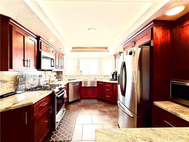 kitchen with light stone countertops, a tray ceiling, stainless steel appliances, dark brown cabinets, and tasteful backsplash