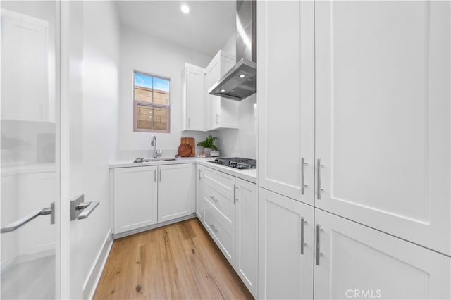 kitchen with a sink, wall chimney range hood, gas stovetop, white cabinets, and light wood finished floors