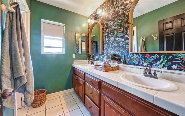 bathroom featuring baseboards, wallpapered walls, double vanity, a sink, and tile patterned flooring