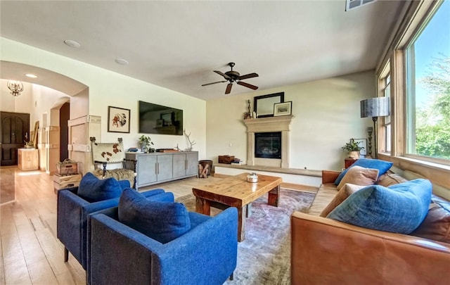 living area featuring a ceiling fan, visible vents, light wood-style flooring, arched walkways, and a glass covered fireplace