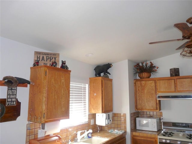 kitchen with tile countertops, gas stove, white microwave, decorative backsplash, and under cabinet range hood