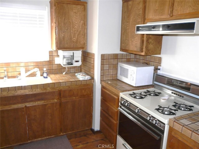 kitchen featuring white appliances, tile counters, under cabinet range hood, brown cabinets, and backsplash