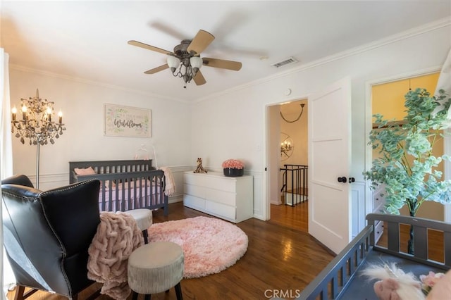 bedroom featuring visible vents, ornamental molding, ceiling fan with notable chandelier, wood finished floors, and a nursery area