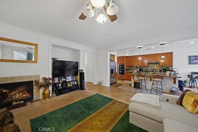 living area featuring a ceiling fan, light tile patterned flooring, a tiled fireplace, crown molding, and light colored carpet