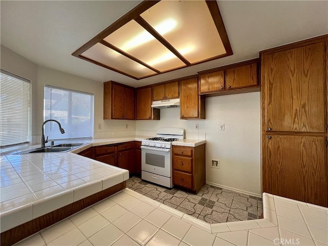 kitchen with brown cabinetry, a sink, tile counters, white gas range oven, and under cabinet range hood