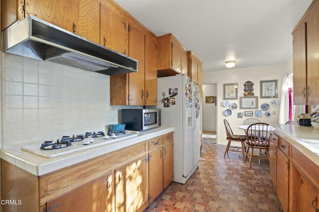 kitchen with brown cabinets, under cabinet range hood, backsplash, tile countertops, and white appliances