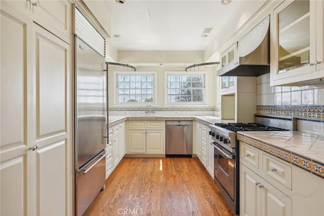 kitchen featuring high quality appliances, light wood-style flooring, under cabinet range hood, tasteful backsplash, and glass insert cabinets