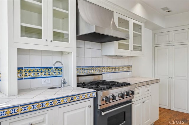 kitchen featuring high end stove, a sink, white cabinetry, wall chimney range hood, and decorative backsplash