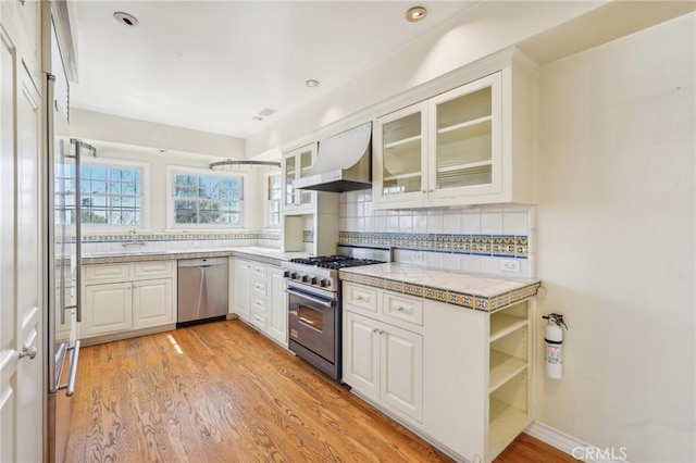 kitchen with light wood-type flooring, backsplash, white cabinetry, appliances with stainless steel finishes, and exhaust hood