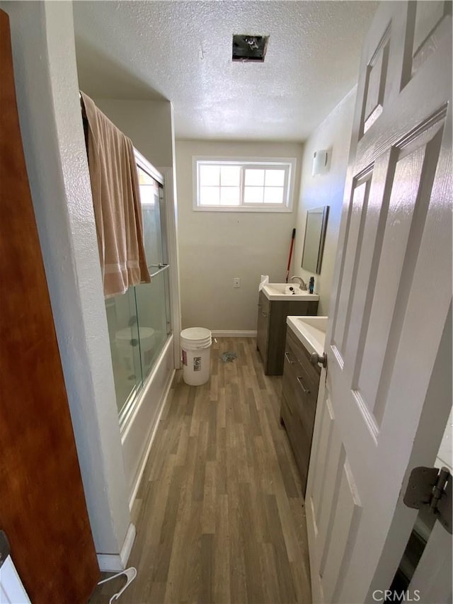 full bath featuring a textured ceiling, wood finished floors, baseboards, bath / shower combo with glass door, and vanity