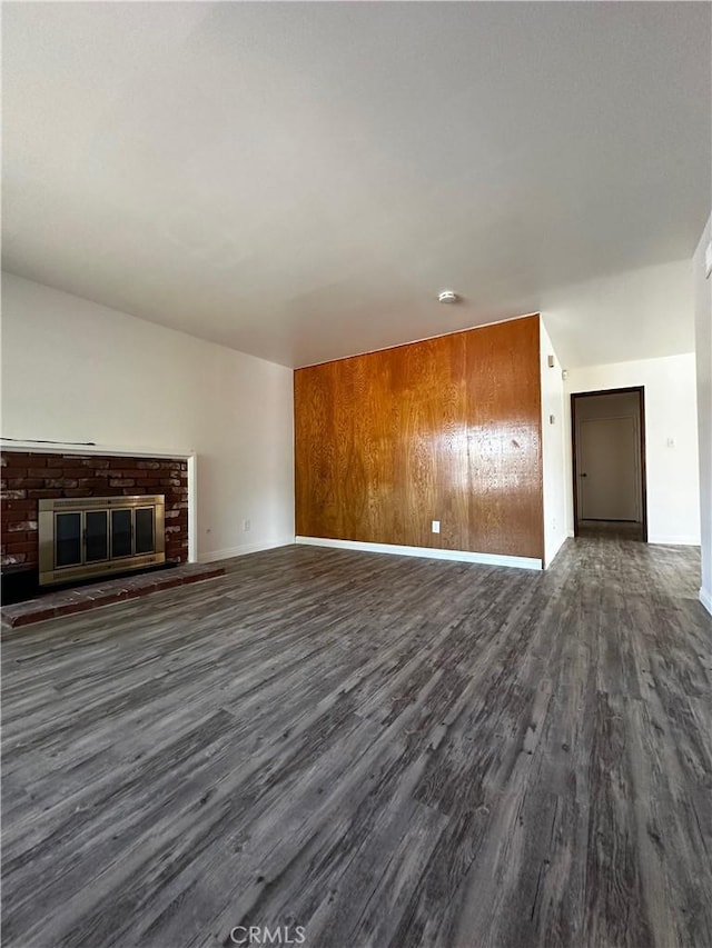 unfurnished living room featuring wood walls, a brick fireplace, baseboards, and dark wood-style flooring