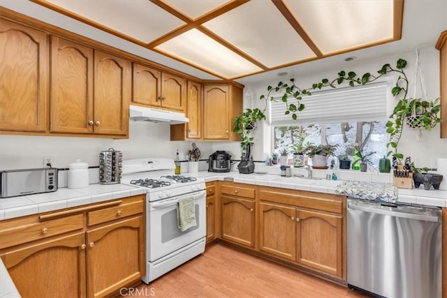 kitchen with under cabinet range hood, white gas stove, light wood-style flooring, brown cabinets, and stainless steel dishwasher