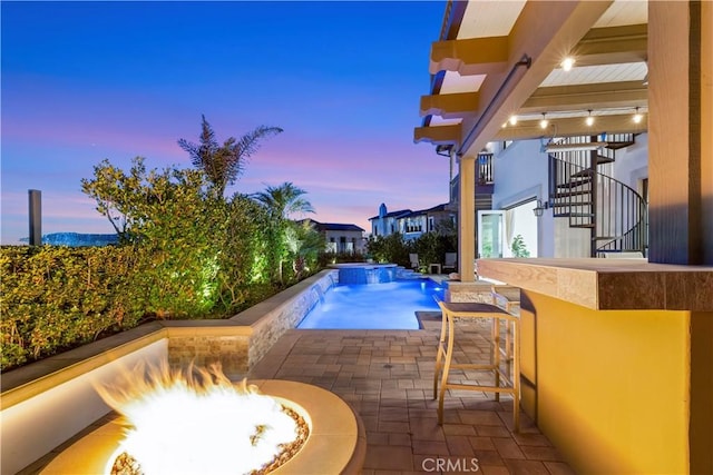 pool at dusk with an outdoor fire pit, stairs, a patio area, outdoor dry bar, and an infinity pool