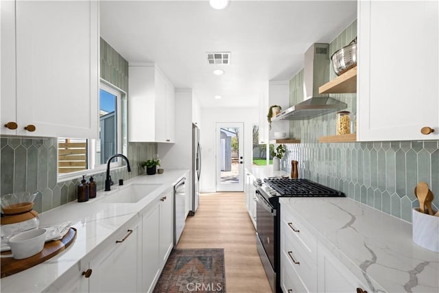 kitchen with visible vents, open shelves, appliances with stainless steel finishes, wall chimney exhaust hood, and a sink