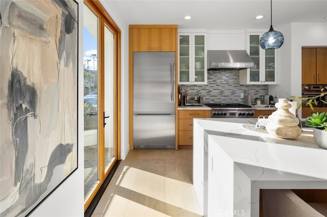 kitchen featuring decorative light fixtures, tasteful backsplash, stainless steel appliances, wall chimney range hood, and glass insert cabinets