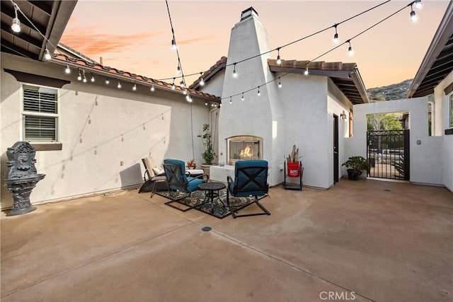 patio terrace at dusk featuring a lit fireplace and a gate